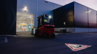 Wózek widłowy Linde wyposażony w system Linde TruckSpot wyjeżdża tyłem z hali o zmierzchu. Wózek widłowy Linde wyposażony w system Linde TruckSpot wyjeżdża tyłem z hali o zmierzchu.