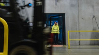 Mężczyzna w odzieży ochronnej wychodzi przez drzwi do hali fabrycznej. Obok przejeżdża wózek widłowy. Mężczyzna w odzieży ochronnej wychodzi przez drzwi do hali fabrycznej. Obok przejeżdża wózek widłowy.
