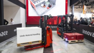 Dwa wózki widłowe Linde MH transportują towary. Cyfrowy obraz wózków widłowych można zobaczyć na ekranie projekcyjnym.