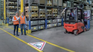 Linde TruckSpot wyświetla symbol na podłodze magazynu Dwie osoby poruszają się po magazynie, a ich trasa krzyżuje się z trasą przejazdu wózka
