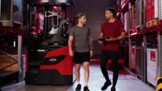 Dwie osoby w strojach sportowych stoją przed wózkiem widłowym wysokiego składowania Ri16 firmy Linde MH; jedna z nich trzyma tablet.