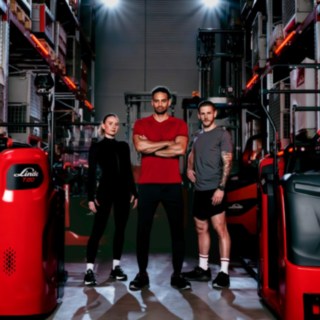Kobieta i dwóch mężczyzn w strojach sportowych stoją w magazynie pośród wózków widłowych firmy Linde Material Handling.