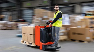 Dzięki funkcji Linde e-driver operatorzy mogą sterować wszystkimi funkcjami wózka jedną ręką, nie obciążając kręgosłupa. Wózek unoszący Linde T20 przejeżdża przez magazyn, operator stoi w pozycji ustawionej pod kątem 45 stopni