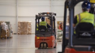 Elektryczny wózek widłowy Linde z operatorem w kamizelce Welcome Airpot Services przewozi paletę w dużym magazynie cargo, obok innych pracowników i wózków